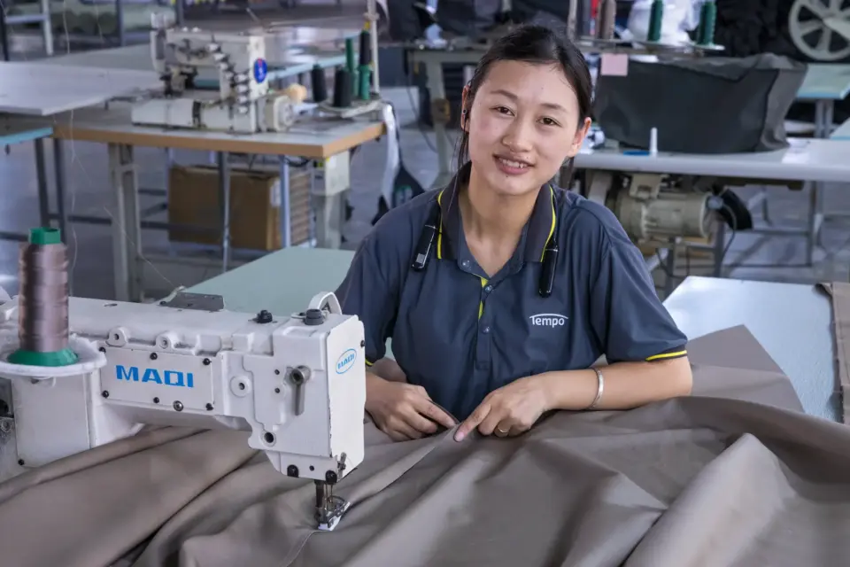 A skilled seamstress wearing a Tempo company polo shirt smiles while operating an industrial sewing machine to expertly stitch a durable outdoor umbrella canopy.